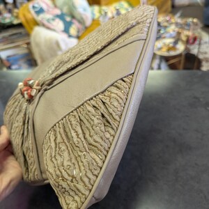 Puede incluir: Un bolso de mano vintage beige con un patr&oacute;n texturizado en forma de onda. El bolso presenta un panel beige liso contrastante y un cierre decorativo con detalles de color coral. El dise&ntilde;o incluye detalles de tela plisada.