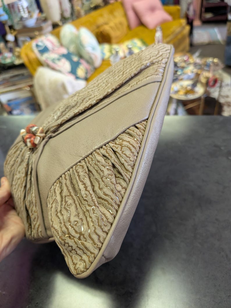 Puede incluir: Un bolso de mano vintage beige con un patr&oacute;n texturizado en forma de onda. El bolso presenta un panel beige liso contrastante y un cierre decorativo con detalles de color coral. El dise&ntilde;o incluye detalles de tela plisada.