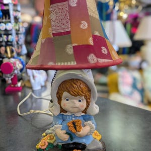 May include: A vintage table lamp featuring a patchwork lampshade in shades of red, orange, and gold. The base is a ceramic figurine of a girl in a blue dress holding a butterfly, surrounded by flowers. The lamp has a white cord.