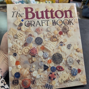 May include: The Button Craft Book by Dawn Cusick. The book cover is filled with a variety of buttons in different shapes, sizes, and colors, including white, cream, blue, red, and black. The title is in a burgundy color.