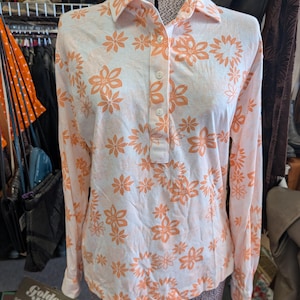 May include: A vintage long-sleeve shirt with a floral pattern. The shirt is white with orange flowers and has a collared neckline and button-up front. The shirt is displayed on a mannequin.