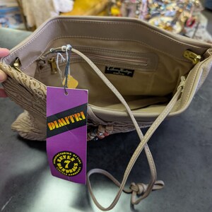 Puede incluir: Bolso de mano de cuero beige con una correa larga. El interior es visible, con un peque&ntilde;o bolsillo y una etiqueta. Una etiqueta morada con el nombre "DIMITRI" est&aacute; adjunta. El bolso tiene herrajes dorados.