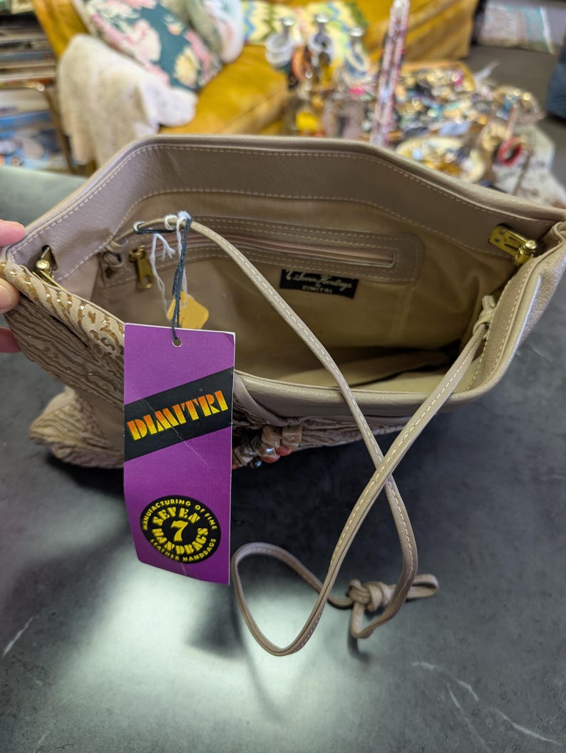 Puede incluir: Bolso de mano de cuero beige con una correa larga. El interior es visible, con un peque&ntilde;o bolsillo y una etiqueta. Una etiqueta morada con el nombre "DIMITRI" est&aacute; adjunta. El bolso tiene herrajes dorados.