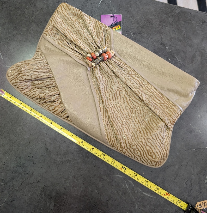 Puede incluir: Bolso de mano beige con solapa texturizada y un acento decorativo de cuentas. El bolso de mano mide aproximadamente 43 cm de largo. La bolsa tiene una etiqueta que dice "TRI".