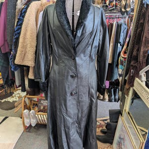 May include: A long, black leather coat with a faux fur collar and cuffs. The coat is buttoned up and displayed on a mannequin. The coat is part of a vintage clothing collection.