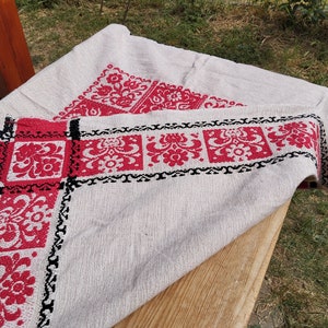 May include: A white linen tablecloth with red and black embroidered floral designs. The tablecloth is folded and draped over a wooden surface.