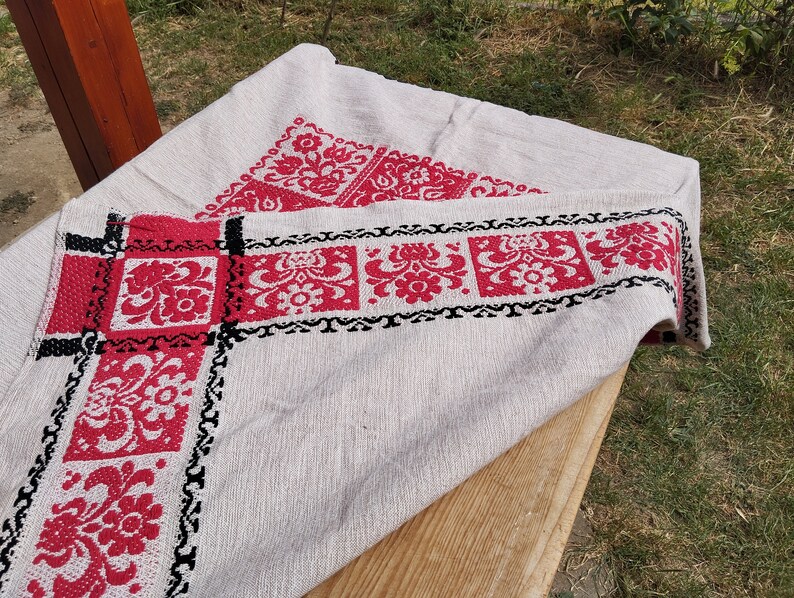 May include: A white linen tablecloth with red and black embroidered floral designs. The tablecloth is folded and draped over a wooden surface.