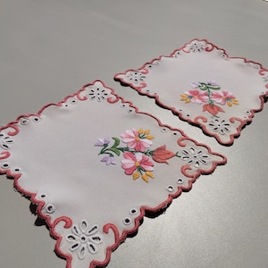 May include: Two white embroidered placemats with pink scalloped edges and floral designs. Each placemat features a central embroidered bouquet of pink, yellow, and purple flowers with green leaves. The edges have cut-out floral patterns.