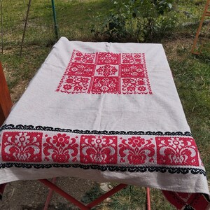 May include: A white tablecloth with a red and black embroidered floral design. The tablecloth is folded and placed on a red metal folding table.