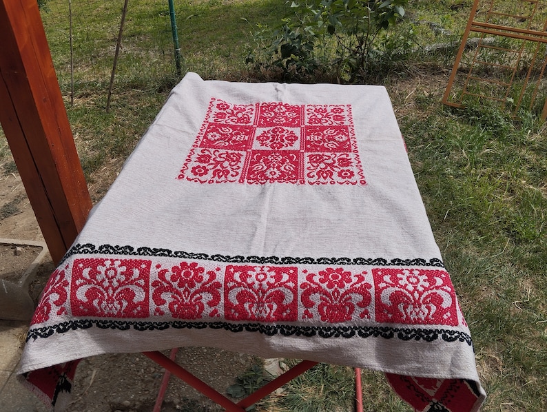 May include: A white tablecloth with a red and black embroidered floral design. The tablecloth is folded and placed on a red metal folding table.