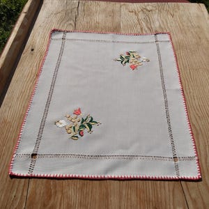 Puede incluir: Un mantel individual rectangular de lino blanquecino con diseños florales bordados. El mantel individual tiene un borde cosido en rojo y esquinas decorativas recortadas. El bordado floral incluye acentos rojos, blancos y verdes.