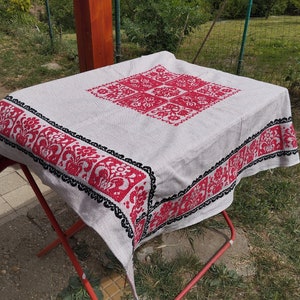 May include: A white tablecloth with a red and black embroidered floral design. The tablecloth is folded and placed on a red metal folding table.