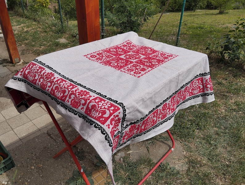 May include: A white tablecloth with a red and black embroidered floral design. The tablecloth is folded and placed on a red metal folding table.