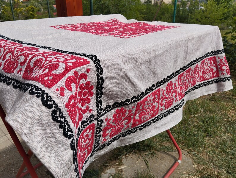 May include: A white tablecloth with a red and black embroidered floral design. The tablecloth is folded and draped over a red metal drying rack.