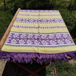 Puede incluir: Textil estampado en amarillo y morado con flecos. El textil tiene un diseño geométrico repetitivo en morado y blanco. Los flecos son de color morado liso. El textil se muestra sobre una mesa roja.