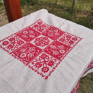 May include: White tablecloth with a red embroidered floral design in the center. The tablecloth has a black and white border.