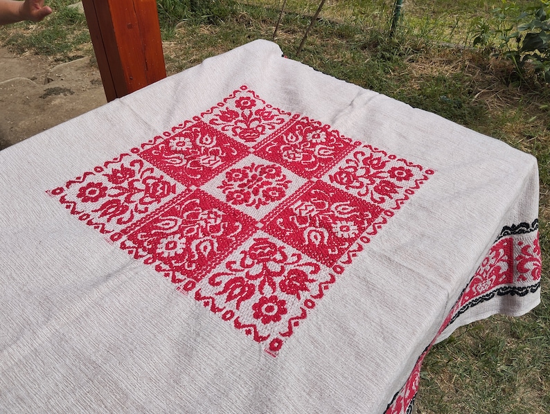 May include: White tablecloth with a red embroidered floral design in the center. The tablecloth has a black and white border.