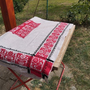 May include: A white linen tablecloth with red and black embroidered floral designs. The tablecloth is folded and laid out on a wooden table.