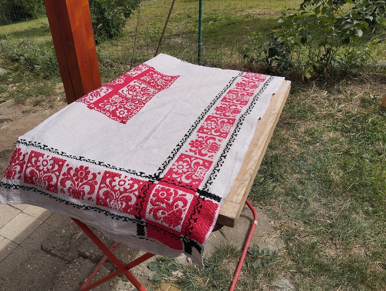 May include: A white linen tablecloth with red and black embroidered floral designs. The tablecloth is folded and laid out on a wooden table.