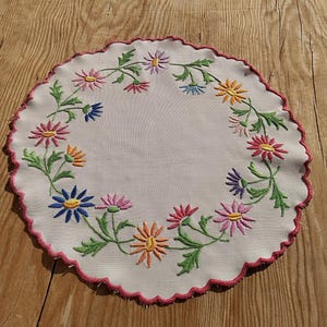 Puede incluir: Un mantel individual circular blanco con un borde festoneado y bordado con una corona de flores de colores.