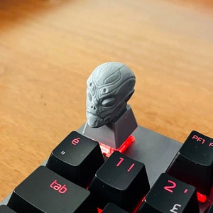 May include: A gray alien head keycap sits atop a black mechanical keyboard. The keycaps have red lettering and symbols. The keyboard is on a wooden surface. The alien head is detailed with facial features.