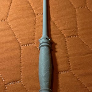 Op de afbeelding: Een lichtgrijze toverstaf met een lange, slanke schacht en een handvat met een textuur van ruitvormige patronen. Het handvat is voorzien van decoratieve, kroonachtige uitlopende ringen aan zowel de bovenkant als de onderkant van de greep.