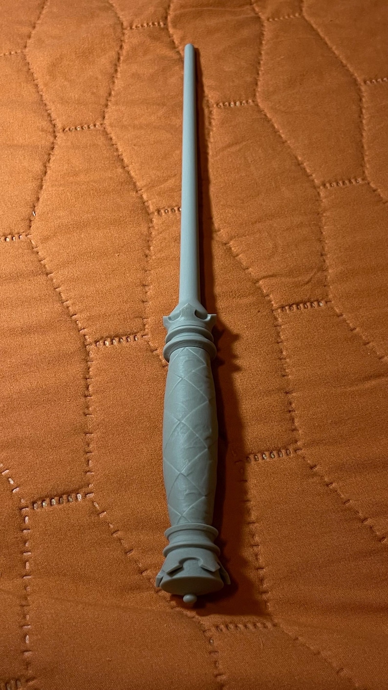 Op de afbeelding: Een lichtgrijze toverstaf met een lange, slanke schacht en een handvat met een textuur van ruitvormige patronen. Het handvat is voorzien van decoratieve, kroonachtige uitlopende ringen aan zowel de bovenkant als de onderkant van de greep.