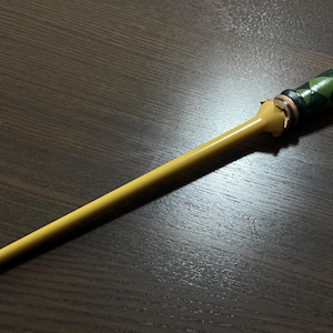 Op de afbeelding: Een lange, slanke toverstaf met een gladde, effen gele schacht en een getextureerd handvat. Het handvat is versierd met een donkergroen en zwart ruitpatroon, geaccentueerd door metalen zilverkleurige ringen aan de boven- en onderkant.