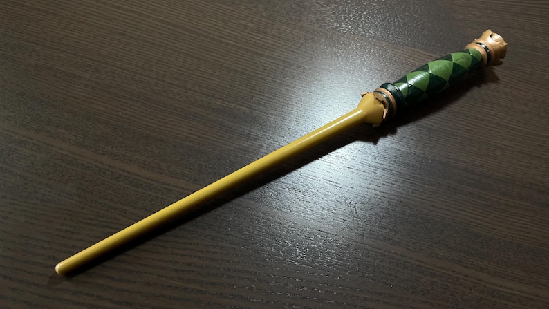 Op de afbeelding: Een lange, slanke toverstaf met een gladde, effen gele schacht en een getextureerd handvat. Het handvat is versierd met een donkergroen en zwart ruitpatroon, geaccentueerd door metalen zilverkleurige ringen aan de boven- en onderkant.