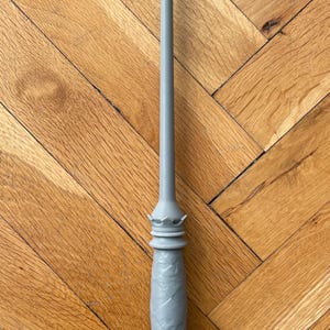 Op de afbeelding: Een lichtgrijze toverstaf met een lange, slanke, taps toelopende schacht en een gestructureerd handvat met een ruitpatroon. Het handvat is voorzien van een kroonachtig detail aan de bovenkant en een uitlopende, ingekeepte basis in &eacute;&eacute;n kleur.