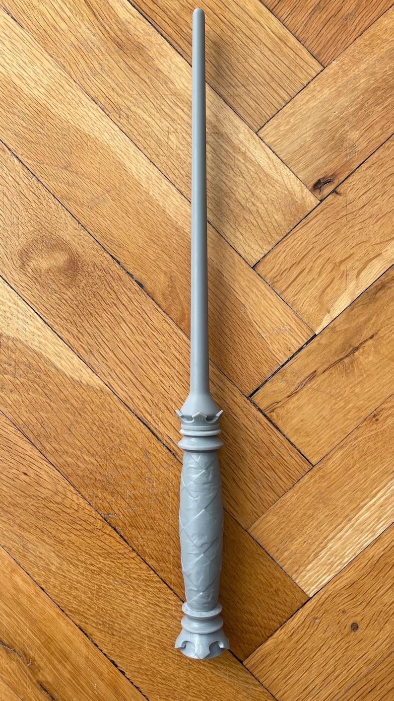 Op de afbeelding: Een lichtgrijze toverstaf met een lange, slanke, taps toelopende schacht en een gestructureerd handvat met een ruitpatroon. Het handvat is voorzien van een kroonachtig detail aan de bovenkant en een uitlopende, ingekeepte basis in &eacute;&eacute;n kleur.