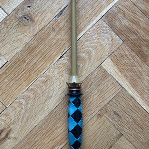 Op de afbeelding: Een toverstaf met een lange, gladde, lichtbruine taps toelopende schacht. Het handvat is versierd met een zwart en lichtblauw ruitpatroon, geaccentueerd door een donkerbruine kroonvormige kraag aan de bovenkant en een bijpassende voet onderaan.
