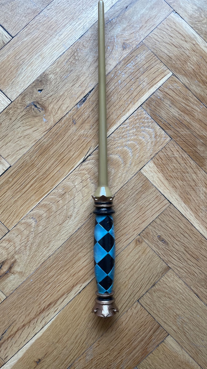 Op de afbeelding: Een toverstaf met een lange, gladde, lichtbruine taps toelopende schacht. Het handvat is versierd met een zwart en lichtblauw ruitpatroon, geaccentueerd door een donkerbruine kroonvormige kraag aan de bovenkant en een bijpassende voet onderaan.