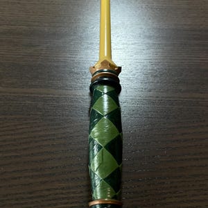 Op de afbeelding: Een decoratieve toverstaf met een lange, gladde, gele, taps toelopende schacht. Het handvat is cilindervormig met een donkergroen en lichtgroen ruitpatroon. Het is geaccentueerd met metallic goudkleurige ringen en een kroonvormige basis en kraag.
