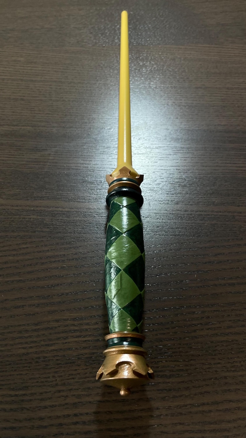 Op de afbeelding: Een decoratieve toverstaf met een lange, gladde, gele, taps toelopende schacht. Het handvat is cilindervormig met een donkergroen en lichtgroen ruitpatroon. Het is geaccentueerd met metallic goudkleurige ringen en een kroonvormige basis en kraag.