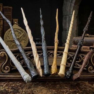 May include: A collection of eight distinct wands displayed in a decorative wooden stand. The wands feature varied textures and finishes, including light grey, dark charcoal, spiral-carved beige, black, natural wood grain, and dark brown wood tones.