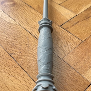 Op de afbeelding: Een grijze kunststof toverstaf met een lange, slanke schacht en een handvat met een textuur van ruitpatronen. Het handvat is aan de boven- en onderkant voorzien van uitlopende, decoratieve ronde ringen en eindigt in een kleine, ronde knop.