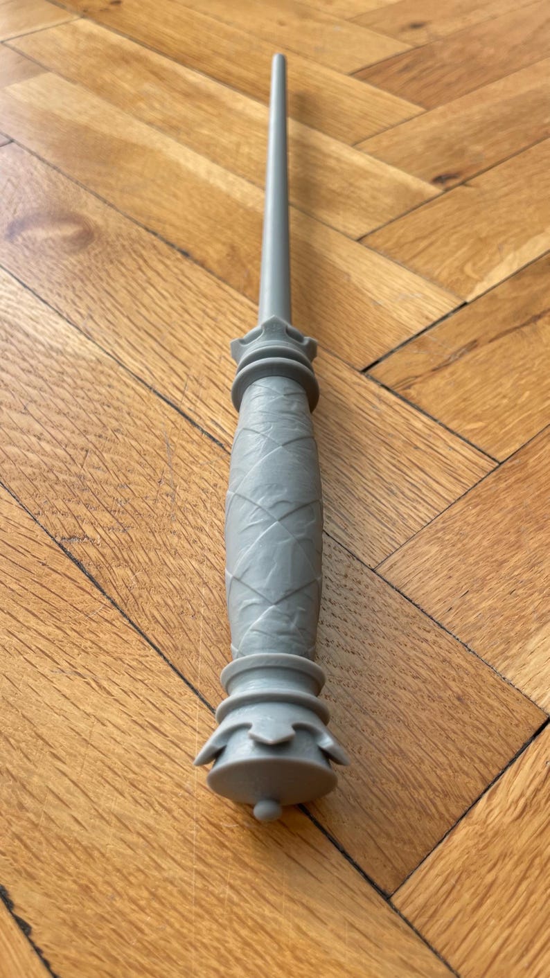 Op de afbeelding: Een grijze kunststof toverstaf met een lange, slanke schacht en een handvat met een textuur van ruitpatronen. Het handvat is aan de boven- en onderkant voorzien van uitlopende, decoratieve ronde ringen en eindigt in een kleine, ronde knop.