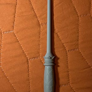 Op de afbeelding: Een lichtgrijze toverstaf met een lange, slanke schacht en een getextureerd handvat. Het handvat heeft een ruitvormig patroon en is afgewerkt met kroonachtige decoratieve elementen aan zowel de bovenkant als de onderkant van de greep.