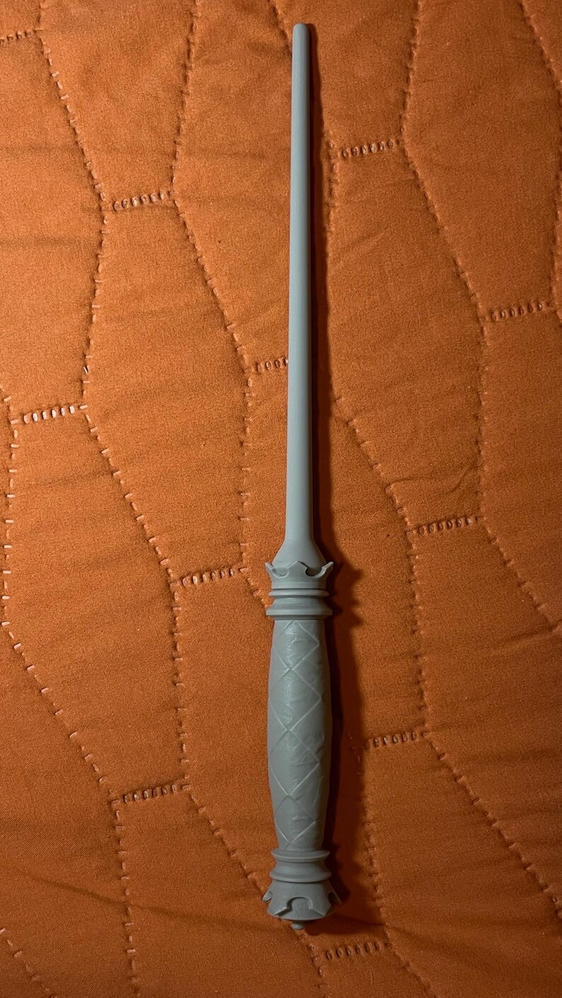 Op de afbeelding: Een lichtgrijze toverstaf met een lange, slanke schacht en een getextureerd handvat. Het handvat heeft een ruitvormig patroon en is afgewerkt met kroonachtige decoratieve elementen aan zowel de bovenkant als de onderkant van de greep.