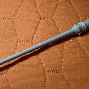 Op de afbeelding: Een lichtgrijze toverstaf met een lange, slanke schacht en een handvat met een textuur van ruitpatronen. Het handvat heeft een uitlopende, geringde kraag bij de basis van de schacht en de knop aan het uiteinde heeft een gekarteld, kroonachtig ontwerp.