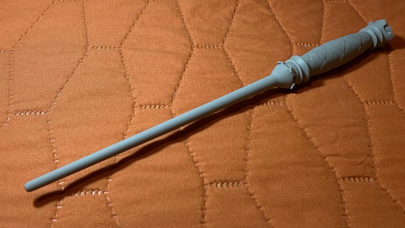 Op de afbeelding: Een lichtgrijze toverstaf met een lange, slanke schacht en een handvat met een textuur van ruitpatronen. Het handvat heeft een uitlopende, geringde kraag bij de basis van de schacht en de knop aan het uiteinde heeft een gekarteld, kroonachtig ontwerp.