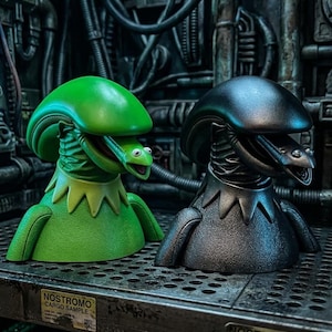 Op de afbeelding: Twee bustefiguren met een hybride wezenontwerp met een gladde, langwerpige kop en een uitstekende binnenkaak. Eén figuur is felgroen en de andere is metallic zwart. Beide tonen de tekst NOSTROMO CARGO SAMPLE.