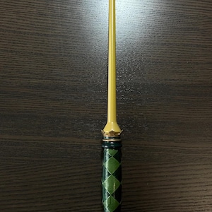 Op de afbeelding: Een slanke toverstaf met een gladde, lichtgele, taps toelopende schacht. Het handvat is versierd met een donker- en lichtgroen ruitpatroon, geaccentueerd door metallic goudkleurige ringen en een kroonachtig detail aan zowel de basis als de top.