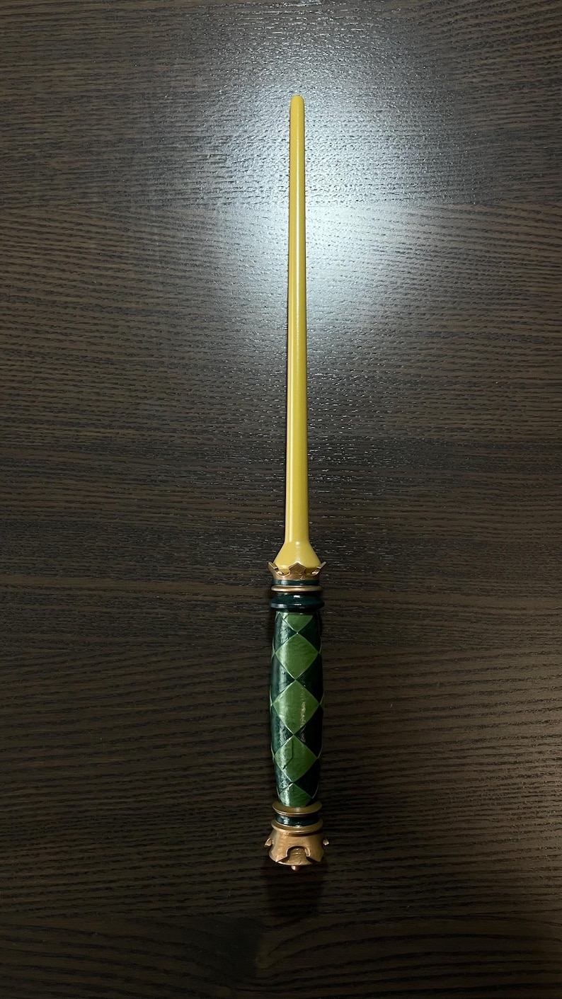 Op de afbeelding: Een slanke toverstaf met een gladde, lichtgele, taps toelopende schacht. Het handvat is versierd met een donker- en lichtgroen ruitpatroon, geaccentueerd door metallic goudkleurige ringen en een kroonachtig detail aan zowel de basis als de top.