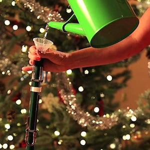 May include: A green watering can pouring water into a clear plastic funnel. The funnel is being held by a hand. The background is a Christmas tree with white lights.