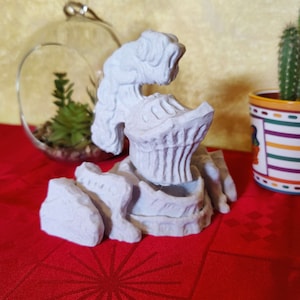 May include: A white 3D printed model of a helmet with a textured surface. The helmet is sitting on a base of rocks, also 3D printed. The model is on a red tablecloth.