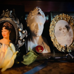 May include: A white plaster bust of a man wearing a laurel wreath, flanked by two oval picture frames containing portraits of women. A small circular red and gold medallion rests in front of the bust, and a pale yellow artificial rose sits nearby.