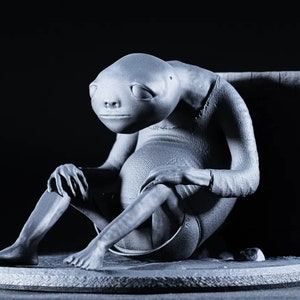 May include: A gray 3D printed figurine of a creature sitting with its legs crossed. The creature has a large head with large eyes and a small mouth. The figurine is sitting on a round base.
