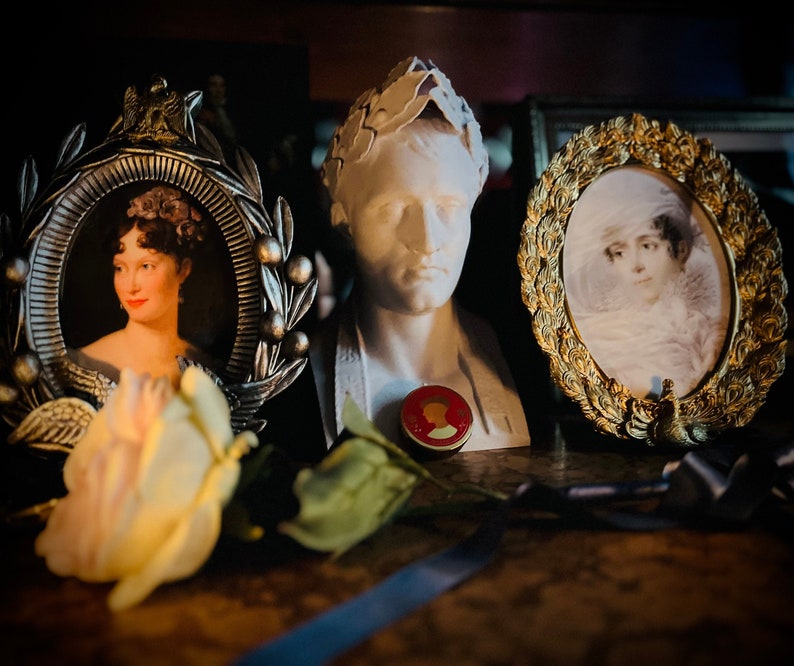 May include: A white plaster bust of a man wearing a laurel wreath, flanked by two oval picture frames containing portraits of women. A small circular red and gold medallion rests in front of the bust, and a pale yellow artificial rose sits nearby.